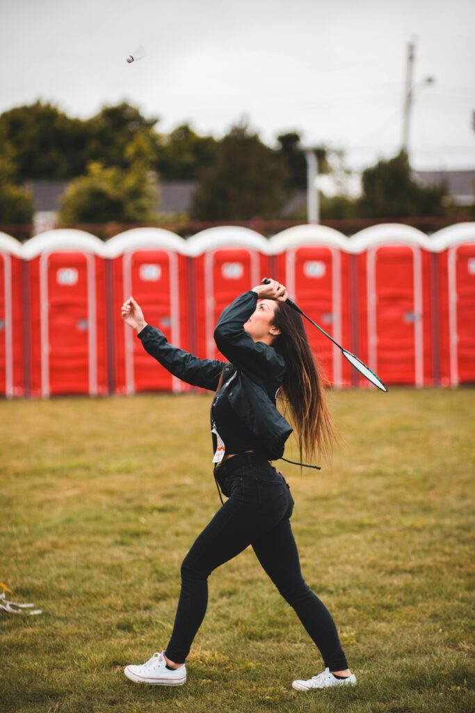 A young woman enjoying a badminton game outdoors with red portable toilets in the background.